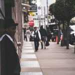A man in a suit and hat leans against a city building, observing pedestrians. — How 2026 Medicare Advantage Plans Compare to