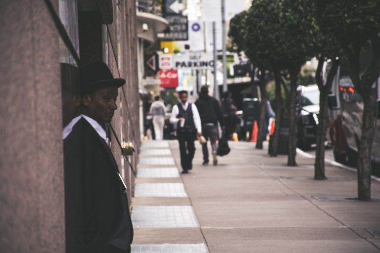 A man in a suit and hat leans against a city building, observing pedestrians. — How 2026 Medicare Advantage Plans Compare to