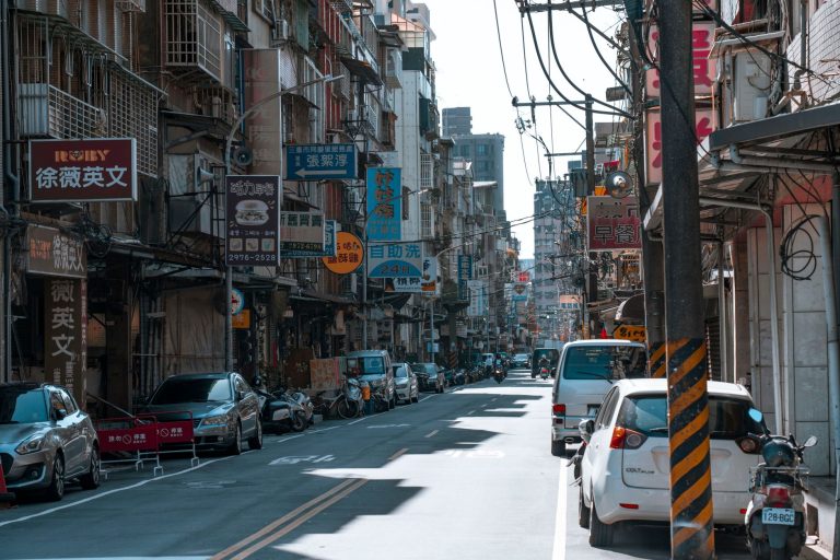 A busy street lined with cars and various colorful business signs on a sunny day. — Urban Oasis of Sensuality: Explore Nuru M